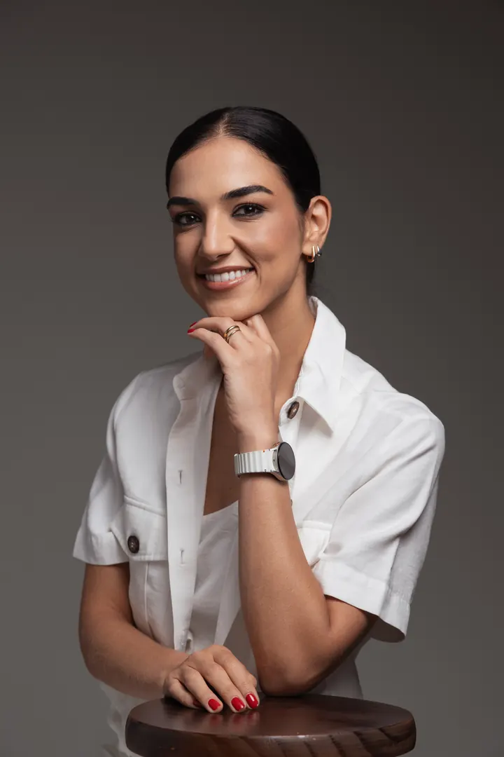 Dra. Luciana Paschoal, geriatra em São Paulo, de camisa branca, apoiada em banqueta de madeira